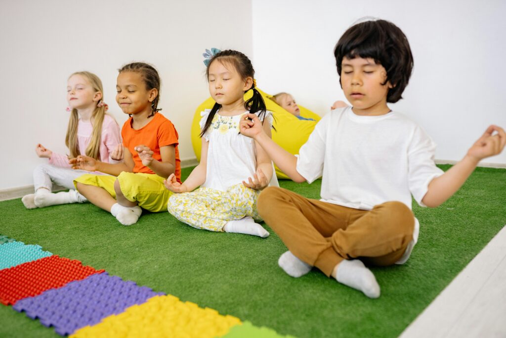 Group of diverse young children meditating in yoga poses on a green mat indoors.